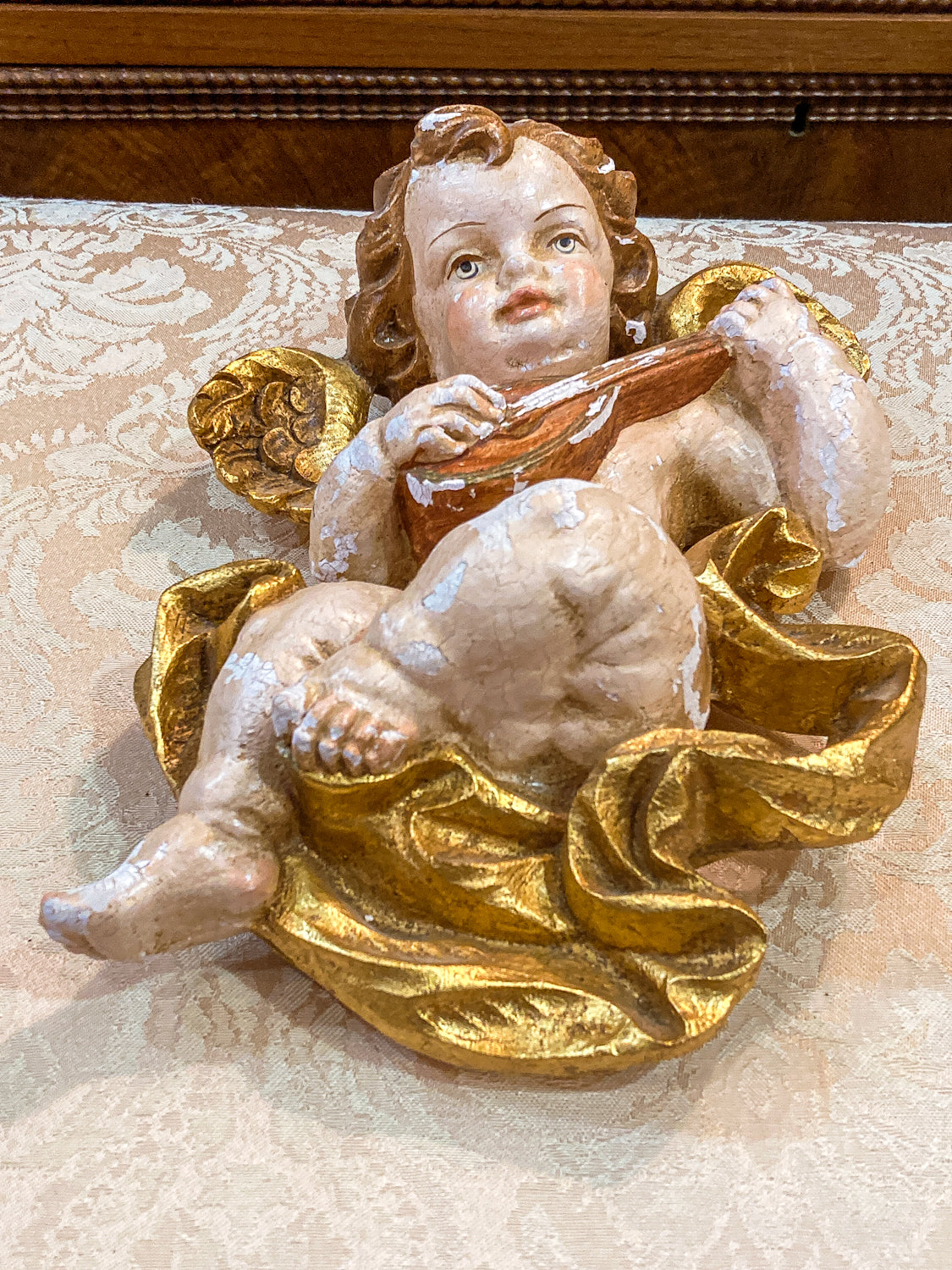 Decorative angel figurine with gold wings and a brown object, on a textured surface. 20th Century Hand Painted Gessoed Gilded Cherub Wall Hanging Sculpture